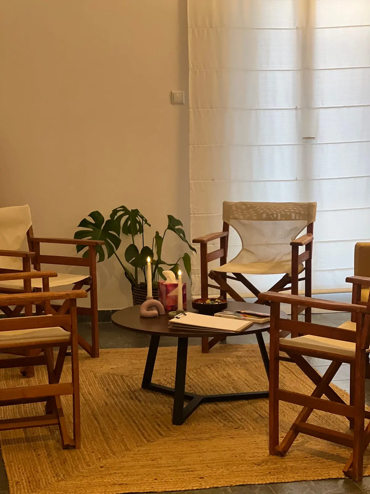 Ξύλινες καρέκλες σκηνοθέτη σε κύκλο γύρω από ένα στρογγυλό τραπεζάκι, πάνω σε ψάθινο χαλί, με μια μονστέρα δίπλα στο παράθυρο